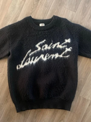 Svart Saint Laurent stickad tröja - Mysig svart stickad tröja från Saint Laurent med fluffig textur och vit logga i graffiti-stil på bröstet. Tröjan har rund halsringning, långa ärmar och ribbade muddar. Perfekt för dig som vill ha en trendig och exklusiv look.