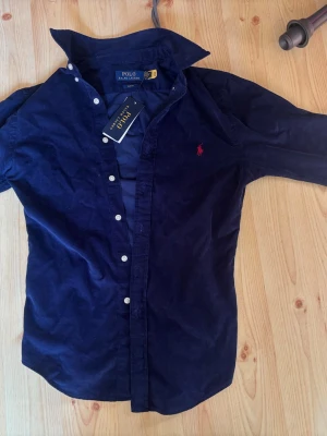Marinblå manchesterskjorta Polo Ralph Lauren - Säljer en marinblå manchesterskjorta från Polo Ralph Lauren med slim fit-passform. Skjortan har klassisk krage, vita knappar och den ikoniska röda logotypen broderad på bröstet. Perfekt för dig som gillar stilrena detaljer och mjukt material.