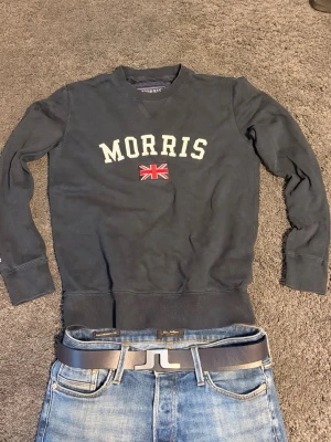 Mörkblå sweatshirt från Morris - Cool mörkblå sweatshirt från Morris med vit text och brittisk flagga på bröstet. Tröjan har rund halsringning, långa ärmar och ribbade muddar. Perfekt för en avslappnad och stilren look.