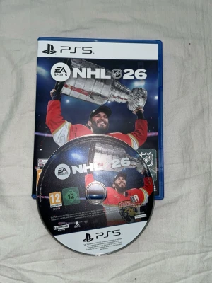 NHL 26 till PS5 - NHL 26 till PlayStation 5 med fodral och skiva. Omslaget har en ishockeyspelare i röd tröja som lyfter Stanley Cup-pokalen. Spelet är ett sportspel med fokus på ishockey och har åldersmärkning 12 år. Perfekt för dig som gillar hockey och gaming.