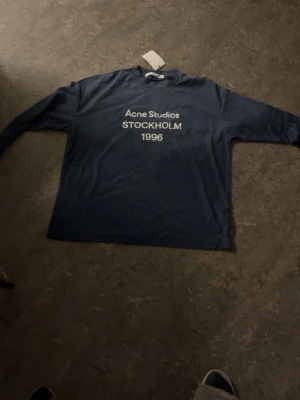 Blå långärmad Acne Studios t-shirt - Snygg mörkblå långärmad t-shirt från Acne Studios med vit text 'STOCKHOLM 1996' på bröstet. Klassisk passform och rund halsringning. Perfekt för dig som gillar stilrena plagg med en urban touch.