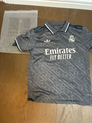 Grå Real Madrid Adidas tröja - Snygg grå Real Madrid tröja från Adidas med korta ärmar och krage. Tröjan har ett diskret mönster, vita detaljer på kragen och ärmsluten samt klubbmärke och Adidas-logga på bröstet. Materialet är troligtvis polyester, perfekt för när du ska ut.