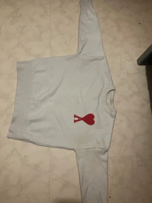 Ljusgrå stickad tröja från Ami - Snygg ljusgrå stickad tröja från Ami Alexandre Mattiussi med röd hjärtlogga och bokstaven A på bröstet. Tröjan har rund hals, ribbade muddar och är tillverkad i mjukt material. Perfekt för dig som gillar stilrena plagg med en twist.