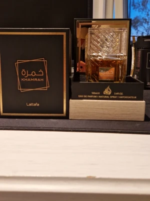Lattafa Khamrah Eau de Parfum 100ml - Lattafa Khamrah Eau de Parfum i en elegant, fyrkantig glasflaska med detaljerat mönster och guldfärgad etikett. Doften kommer i en lyxig svart och guld förpackning. Flaskan rymmer 100 ml och har en exklusiv känsla.