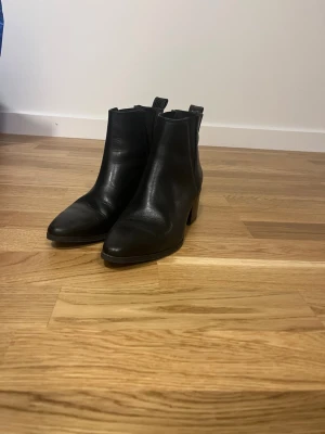Svarta Chelsea boots från Nilson - Snygga svarta Chelsea boots från Nilson i storlek 36. Skorna är gjorda i slätt läder med klassisk resår på sidorna och har en låg klack. Perfekta för dig som vill ha en stilren och tidlös look till vardagsoutfiten.