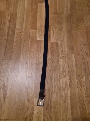 Mörkblått flätat bälte med läderdetalj - Snyggt mörkblått flätat bälte med silverfärgat spänne och bruna läderdetaljer vid spännet. Perfekt för att ge en stilren touch till jeans eller chinos. Bältet har en klassisk design och passar till många olika outfits.