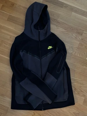 Svart och grå Nike hoodie med dragkedja - Nike hoodie i svart och mörkgrått med hel dragkedja och huva. Snygg V-design på bröstet och en neongul Nike-logga på vänster sida. Fickor på sidorna och en extra ficka på ärmen. Perfekt för en sportig och avslappnad stil.