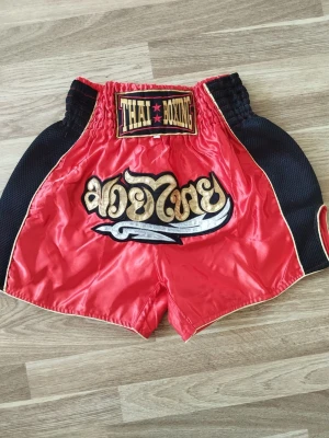 Muay Thai shorts - Köpa i thailand Snygga Muay Thai shorts i röd satin med svarta sidopaneler och guldfärgade detaljer. Resår i midjan för skön passform och broderad text framtill. Perfekta för thaiboxningsträning eller match.