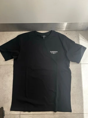 Svart t-shirt med Sardinia-tryck - Säljer en svart t-shirt med vitt tryck på bröstet där det står 'SARDINIA'. T-shirten har rund hals och korta ärmar. Enkel och stilren design som passar till det mesta. Tillverkad i mjuk bomull för skön känsla.finns i många storlekar 