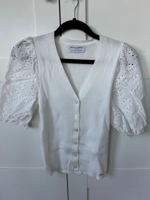 Vit blus med puffärm och spetsdetaljer - Supersöt vit blus från Holly & White med ribbad kropp, v-ringning och knappar framtill. Puffärmarna är i broderad spets med fina hålmönster som ger en romantisk vibe. Perfekt till jeans eller kjol för en fräsch look.