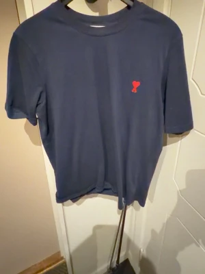 Mörkblå t-shirt från Ami - Säljer en mörkblå t-shirt från Ami Alexandre Mattiussi med ett litet rött hjärta broderat på bröstet. Klassisk passform och rund halsringning. Perfekt till jeans eller shorts för en clean look. Obs kvitto finns ej då den e köpt utomlands