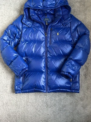 Blå dunjacka från Polo Ralph Lauren - Snygg blå dunjacka från Polo Ralph Lauren med blank finish och gul broderad logga på bröstet. Jackan har huva, dragkedja framtill, två sidofickor med dragkedja och en bröstficka. Perfekt för kalla vinterdagar och riktigt skön att bära. Har bara använt den typ 5 gånger.  Den passar dig mellan 170-180 