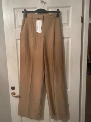 Beige vida kostymbyxor - Säljer ett par beige kostymbyxor med hög midja och vida ben. Byxorna har pressveck framtill, bälteshällor och stängs med dragkedja och knapp. Materialet ser ut att vara en mjuk och lätt tygblandning som ger ett snyggt fall.