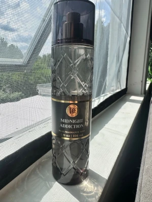 Midnight Addiction Fragrance Mist - Midnight Addiction Fine Fragrance Mist i en elegant, genomskinlig plastflaska med diamantmönster och svart etikett med guldiga detaljer. Doften har inslag av plommon, mandelblomma och tonkaböna. Flaskan rymmer 236 ml och har en praktisk sprayfunktion.