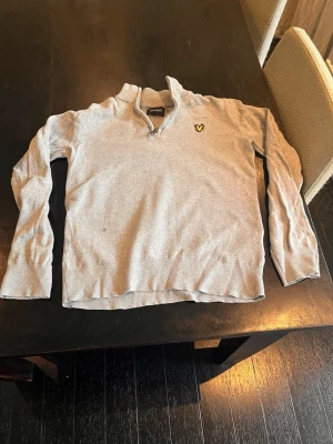 Grå halvzip Lyle & Scott Junior - Snygg grå tröja från Lyle & Scott Junior med halv dragkedja och klassisk gul logga på bröstet. Tröjan har långa ärmar och ribbade muddar. Perfekt för en avslappnad men ändå stilren look.