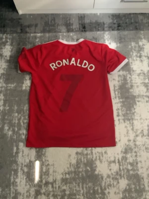 Manchester United Ronaldo matchtröja - Röd Manchester United matchtröja med vita detaljer, Ronaldo och nummer 7 på ryggen. Adidas-logga och klubbmärke på bröstet samt TeamViewer sponsortryck. Klassisk rund hals och korta ärmar, perfekt för fotboll eller supporterliv.