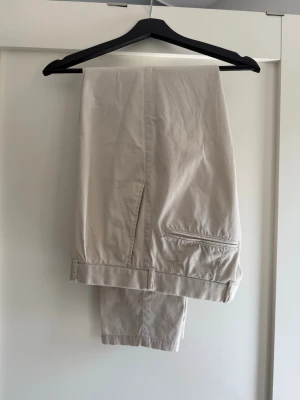 Beige chinos med raka ben - Snygga beige chinos med raka ben och klassisk passform. Byxorna har sidofickor och bakficka med knappdetalj. Tillverkade i ett mjukt och lätt bomullsmaterial som passar perfekt till en avslappnad stil.