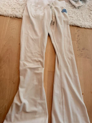 Beiga mjukisbyxor Juicy Couture XS - Säljer ett par beiga mjukisbyxor från Juicy Couture i velour med utsvängda ben och dragsko i midjan. Byxorna har en liten blå logga broderad framtill. Perfekta för en chill och trendig look.