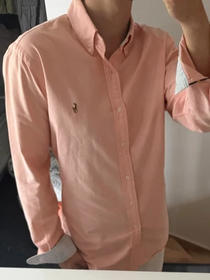 Ljusrosa skjorta från Ralph Lauren - Säljer en klassisk ljusrosa skjorta från Ralph Lauren med button-down krage och broderad logga på bröstet. Skjortan har långa ärmar och vita knappar framtill. Perfekt för dig som vill ha en stilren och tidlös look.