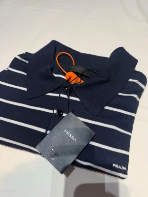 Marinblå randig pikétröja från Prada - Snygg marinblå pikétröja från Prada med vita horisontella ränder och klassisk krage. Tröjan har knäppning framtill med tre knappar och diskret Prada-logga broderad på bröstet. Perfekt för en stilren och trendig look.