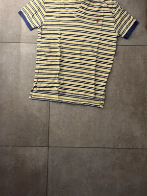 Randig gul och blå polotopp från Polo Ralph Lauren - Snygg polotopp med gula och blå ränder från Polo Ralph Lauren. Klassisk pikékrage, korta ärmar och liten röd logga broderad på bröstet. Perfekt för dig som gillar en sportig och fräsch stil.