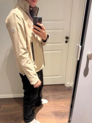 Beige vindjacka från Moncler - Stilren beige vindjacka från Moncler med hög krage och dragkedja framtill. Jackan har två fickor med dragkedja och Moncler-logga på ena ärmen. Perfekt för dig som gillar en clean och sportig look.