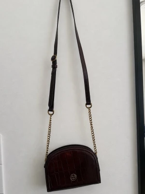 Burgundy axelväska med kedja - Massimo Dutti - Snygg vinröd axelväska i croco-mönstrat skinn med guldfärgad kedja och justerbart axelband. Det finns en mindre, synlig missfärgning/fläck på framsidan, upptill på väskan. Den syns på bilden och är en del av väskans skick, men påverkar inte funktionen eller användningen. I övrigt är väskan i fint skick.