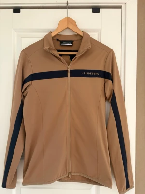 Beige brun ziptröja från J.Lindeberg - Snygg beige brun ziptröja från J.Lindeberg med marinblå rand över bröstet och längs ärmarna. Tröjan har hel dragkedja framtill och står för en sportig och clean look. Perfekt för dig som gillar stilrena plagg med diskreta detaljer.