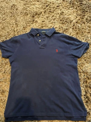 Marinblå pikétröja från Ralph Lauren - Klassisk marinblå pikétröja från Polo Ralph Lauren i bomull. Tröjan har korta ärmar, krage med två knappar och den ikoniska röda logotypen broderad på bröstet. Modellen är slim fit och passar perfekt till jeans eller shorts.