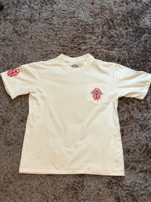 Vit Chrome Hearts t-shirt med tryck - Säljer en riktigt fet vit t-shirt från Chrome Hearts! Kommer med bröstficka, ikoniska Dagger/Vine-grafiken i rött på rygg, ficka och ärm, samt klassiska trycket i nacken.