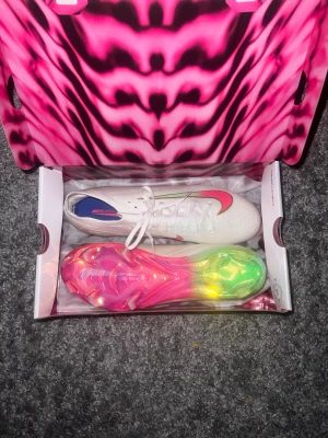 Nike Mercurial vita fotbollsskor - Säljer ett par Nike Mercurial fotbollsskor i vitt med rosa och neongröna detaljer. Skorna har en snygg sula i regnbågsfärger och klassisk Nike Swoosh på sidan. Levereras med originalkartong och en grå Nike-påse. Perfekta för dig som vill sticka ut på planen.