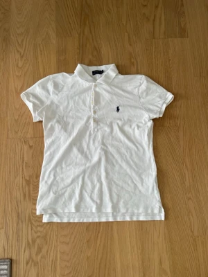 Vit pikétröja från Polo Ralph Lauren - Klassisk vit pikétröja från Polo Ralph Lauren med korta ärmar, krage och knappar framtill. Tröjan har den ikoniska loggan broderad på bröstet och är tillverkad i bomull för en bekväm känsla. Den är i storlek L men passar till 164. Perfekt till jeans eller kjol för en clean look.