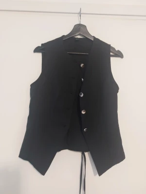 Svart väst med knappar och snörning - Stilren svart väst med knappar framtill och snörning i ryggen. Västen har en klassisk passform och spetsiga kanter nedtill, perfekt för att lyfta din outfit med en edgy touch.Helt ny.