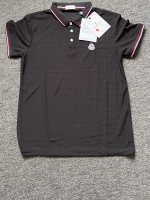 Svart Moncler pikétröja med röd/vit kant - Snygg svart pikétröja från Moncler med klassisk krage och tre knappar framtill. Kragen och ärmsluten har röd och vit kant. Moncler-logga broderad på bröstet. Perfekt till en stilren och sportig look.