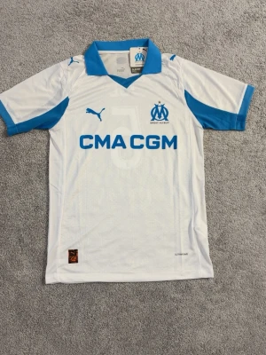 Marseille matchtröja Puma vit/blå - Säljer en vit och blå matchtröja från Olympique de Marseille med nummer 5 och namnet JUL på ryggen. Tröjan har klubbmärke och Puma-logga på bröstet samt sponsortryck framtill. Tillverkad i lätt och ventilerande material, perfekt för fotbollsträning.