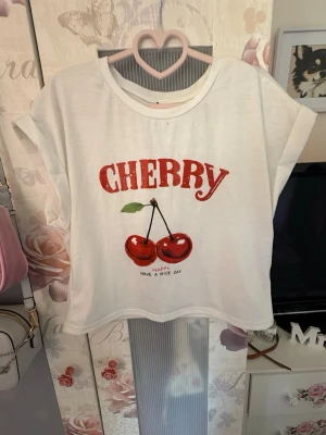 Vit t-shirt med körsbär från SHEIN - Söt vit t-shirt från SHEIN med rött körsbärstryck och texten 'CHERRY' framtill. T-shirten har korta ärmar, rund halsringning och en loose passform. Perfekt för dig som gillar gulliga och färgglada prints.