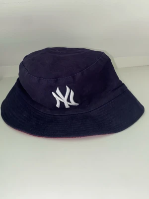 Vändbar bucket hat NY Yankees - Säljer en vändbar bucket hat med New York Yankees-logga. Ena sidan är svart med vit brodyr, andra sidan är röd med svart brodyr. Klassisk form och tillverkad i mjukt tyg, perfekt för en sportig och avslappnad stil.
