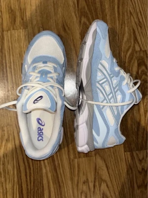 Asics sneakers i blått och vitt - Snygga Asics sneakers i ljusblått och vitt med klassiska ränder på sidan. Ovandelen är i mesh och mockaliknande material för bra andningsförmåga och komfort. Tjock sula i vitt och grått ger en sportig vibe. Perfekta för dig som gillar stilrena och fräscha sneakers.