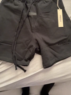Svarta shorts från Essentials - Säljer ett par svarta shorts från Essentials Fear of God med snörning i midjan och diskret logga på benet. Shortsen har en avslappnad passform och är tillverkade i mjuk bomull, perfekta för chill dagar eller streetwear-stil.