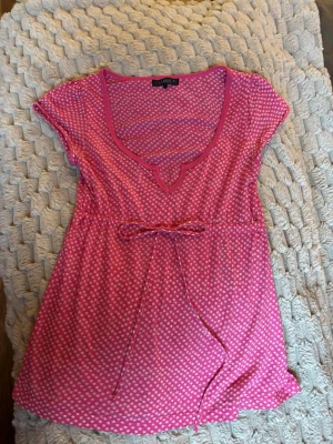 Rosa prickig babydoll topp med knyt - Rosa topp med vita prickar, v-ringad hals och dekorativ knytning i midjan. Kortärmad och i ett lätt, mjukt material som känns skönt mot huden. Perfekt för dig som gillar färg och mönster.