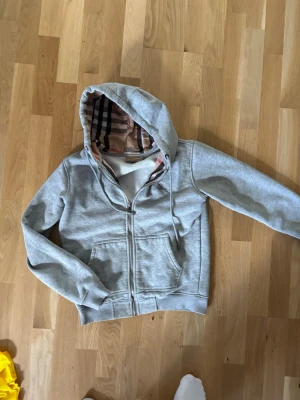 Grå hoodie med dragkedja och huva - Snygg grå hoodie burberry med dragkedja och fickor framtill. Huvan har ett klassiskt rutigt foder i beige, svart och rött som ger en unik touch. Tillverkad i mjukt sweatshirtmaterial och har dragsko i huvan. Perfekt för en chill och avslappnad stil.