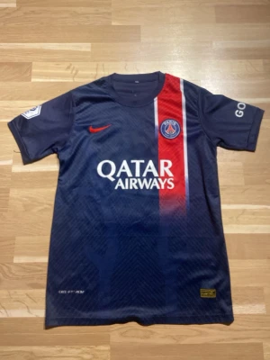 PSG matchtröja Mbappé #7 - Mörkblå PSG matchtröja med röd och vit rand framtill, Qatar Airways-tryck och Nike-logga. På ryggen står det Mbappé och nummer 7. Korta ärmar och officiella klubbmärken på bröst och ärm. Perfekt för fotbollsfans!