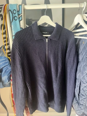 Mörkblå ribbstickad ziptröja från Massimo Dutti - Mörkblå ribbstickad tröja från Massimo Dutti med dragkedja framtill och klassisk krage. Långärmad modell i ett mjukt, tjockare material som passar perfekt för kyligare dagar. Enkel och stilren design som funkar till det mesta.