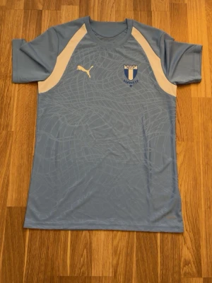 Ljusblå Malmö FF matchtröja Puma - Säljer en ljusblå Malmö FF matchtröja från Puma med vita detaljer på axlarna och klubbmärke på bröstet. Tillverkad i lätt och ventilerande material, perfekt för träning eller match. Snygg diskret mönstring i tyget och klassisk sportig passform.