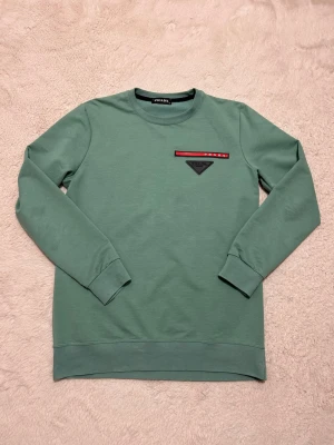 Prada tröja - Snygg grön långärmad topp från Prada med rund halsringning. På bröstet finns en svart och röd detalj med Prada-logga och en svart trekantig patch. Perfekt för dig som gillar stilrena och exklusiva plagg.