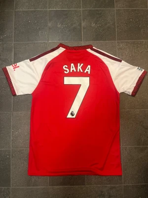 Arsenal matchtröja SAKA #7 - Snygg Arsenal matchtröja i rött och vitt från Adidas. Tröjan har klubbmärke och sponsortryck på bröstet, samt detaljerade ränder på axlarna med Bukayo Saka #7 på ryggen. Tillverkad i ett lätt och ventilerande material, perfekt för fotboll eller träning.