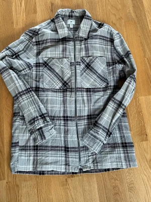 Grå-rutig overshirt med dragkedja - Säljer en stilren overshirt i grått och svart rutmönster. Skjortjackan har två stora bröstfickor, dragkedja framtill och klassisk krage. Tillverkad i mjukt bomullstyg, perfekt för lager-på-lager och en avslappnad look.
