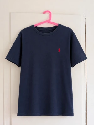 Polo Ralph Lauren marinblå T-shirt 🐎 - Klassisk marinblå T-shirt från Polo Ralph Lauren med den ikoniska röda broderade loggan på bröstet. Stilren och tidlös modell som är lätt att matcha med jeans, shorts eller chinos. Storlek: [fyll i] Skick: Fint skick, se bilder ✨ Skriv gärna vid frågor eller om du vill ha mått!