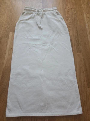 Vit långkjol med snörning - Vit långkjol i mjukt material med elastisk midja och snörning framtill. Kjolen har en rak passform och är enkel att matcha till olika stilar. Perfekt för dig som gillar en clean och avslappnad look.