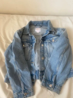 Ljusblå klassisk jeansjacka aldrig använd - Snygg ljusblå jeansjacka med raka linjer och silverfärgade knappar. Jackan har två bröstfickor med lock och knapp samt klassisk krage. Perfekt att slänga över en hoodie eller t-shirt för en avslappnad look.